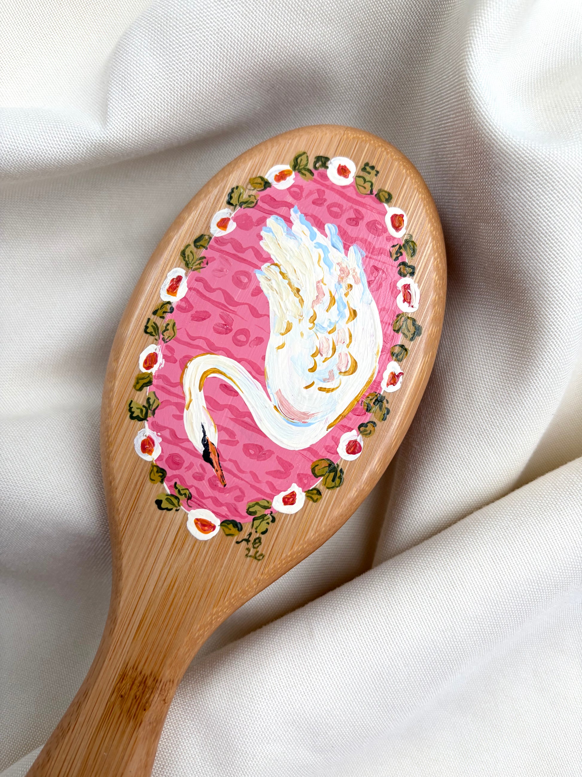 A wooden hairbrush with a hand-painted pink swan design, resting on draped white fabric.