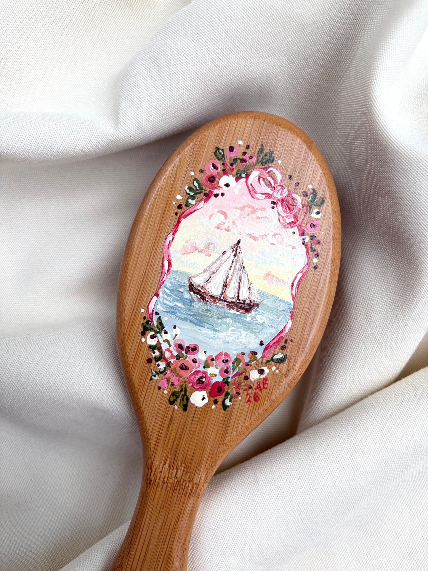 A wooden hairbrush with a hand-painted illustration of a sailboat on the brush head, surrounded by pink and white flowers, resting on draped white fabric.