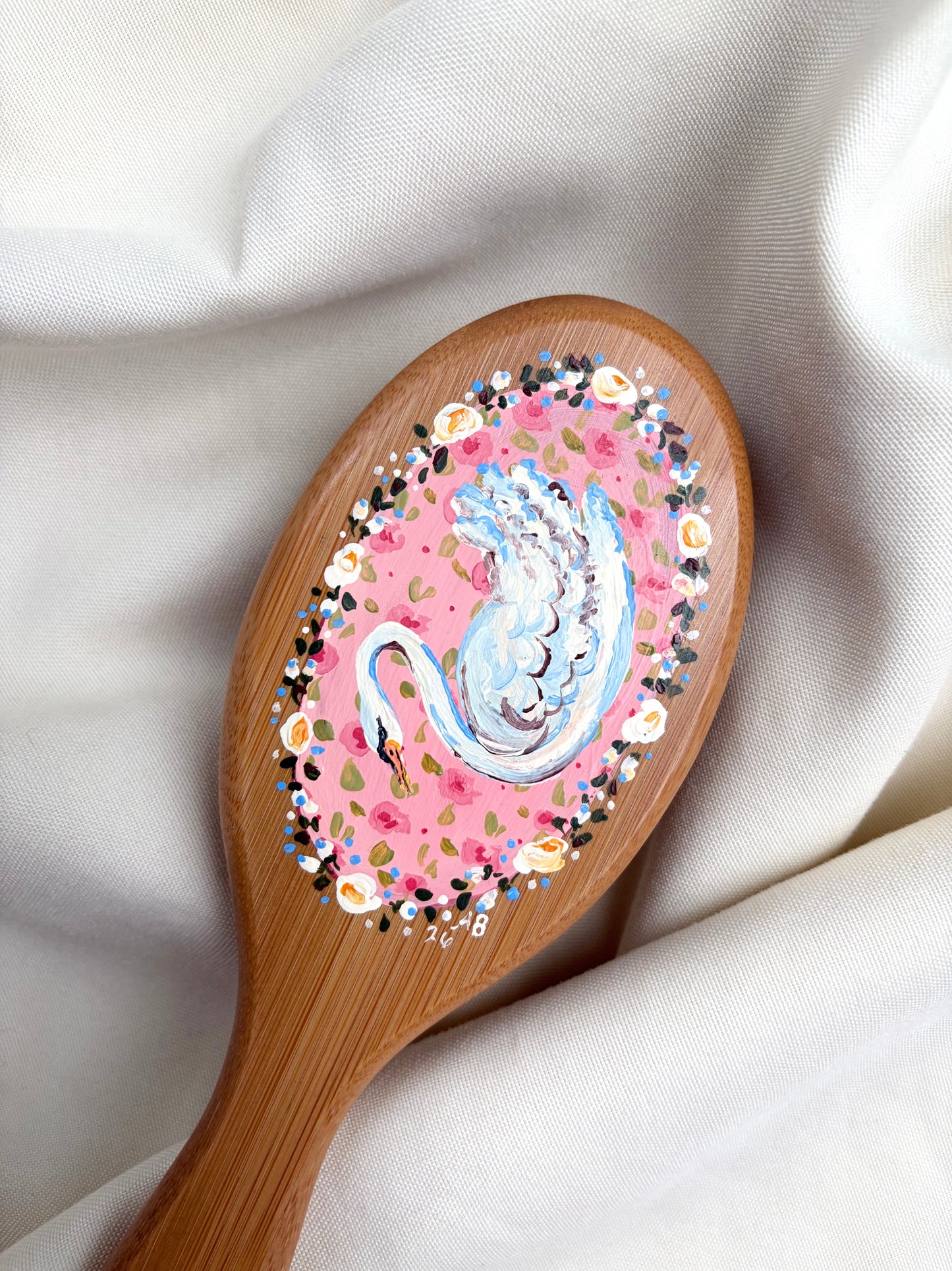 A wooden hairbrush with a hand-painted swan design on the bristle cover, resting on draped white fabric.