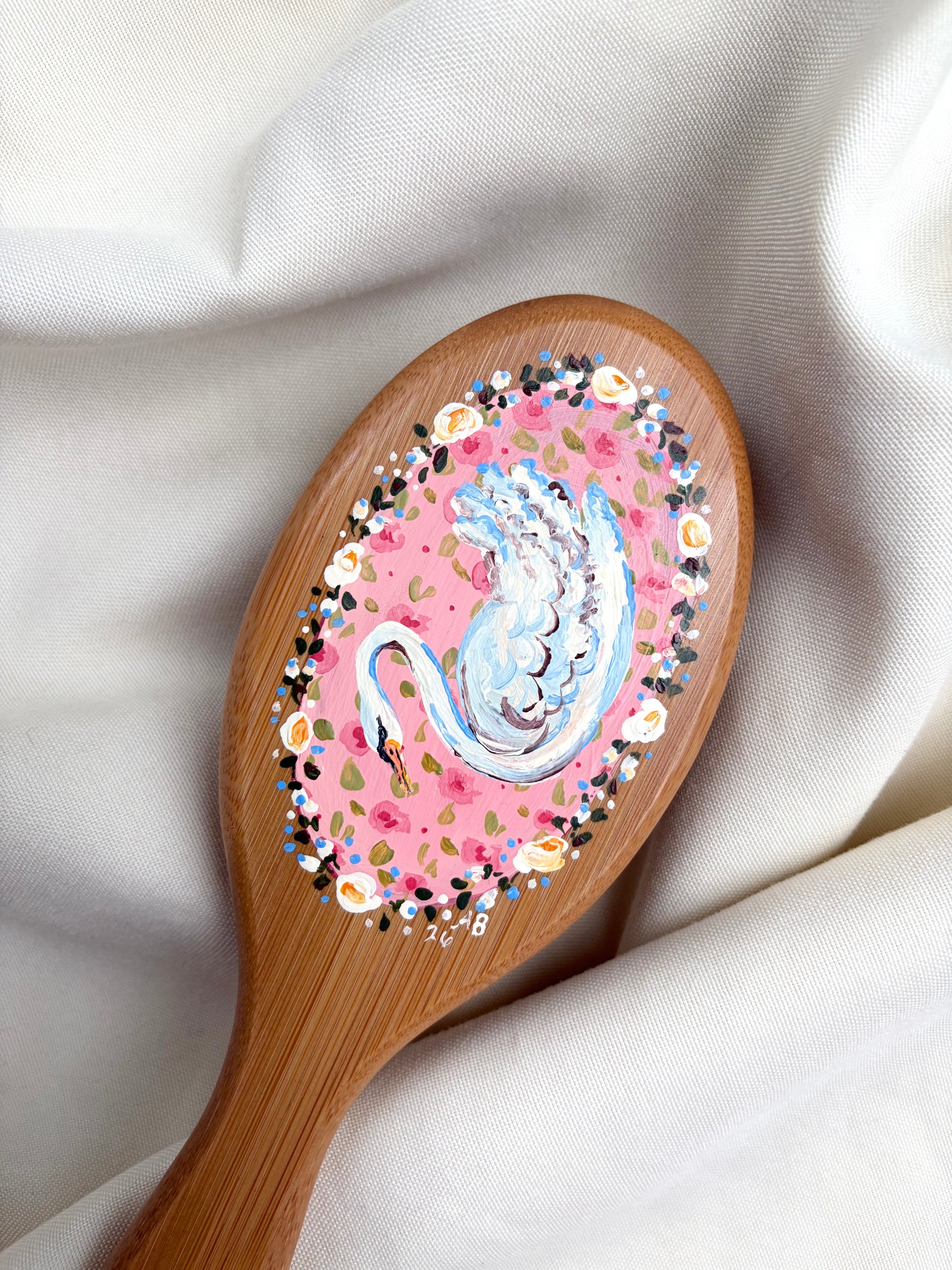 A wooden hairbrush with a hand-painted swan design on the bristle cover, resting on draped white fabric.